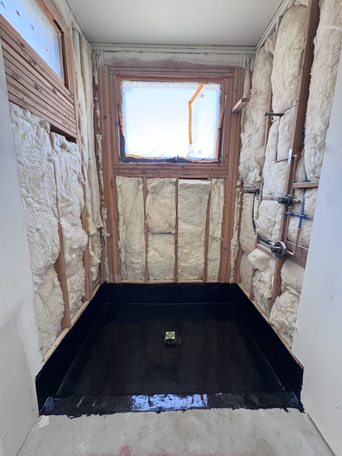 Shower preparation with a waterproofed pan, exposed wall framing, insulation, and rough-in plumbing